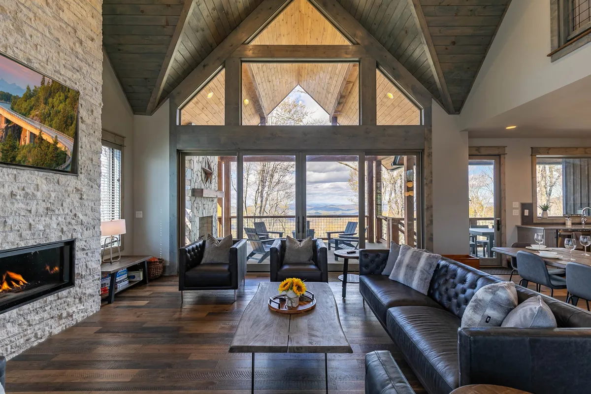 This image showcases a beautiful open-plan living and dining area with large windows framing sweeping mountain views. The space features a comfortable seating area, a modern fireplace, and an adjacent dining table, all bathed in natural light.