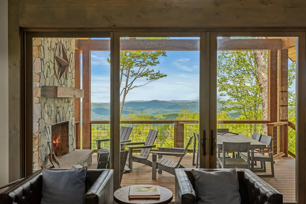 An inviting outdoor wooden deck featuring a stone fireplace, comfortable seating, and sweeping mountain views. The space is ideal for relaxing by the fire, enjoying coffee at sunrise or sunset, and socializing with friends while taking in the natural beauty of the surrounding landscape.
