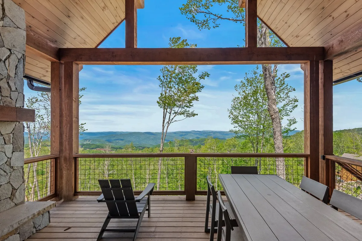 A spacious wooden deck with a dining table and black chairs overlooks the mountains, framed by a stone wall. The tranquil scene suggests outdoor dining, relaxation, and nature watching, aligning well with community events and wine tastings.