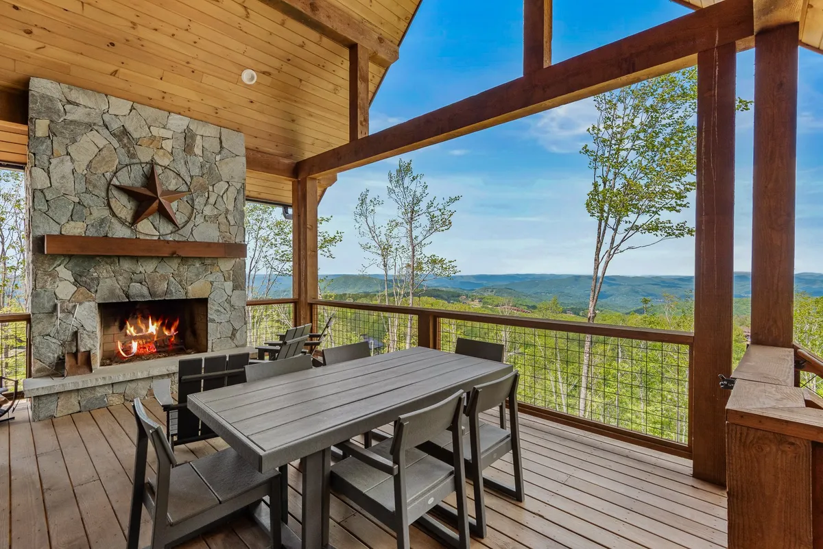 An inviting exterior deck with a stone fireplace, outdoor dining table, and sweeping panoramic mountain view. The setting is ideal for morning coffee, outdoor meals, or cozy evenings gathered around the fireplace under the covered porch.