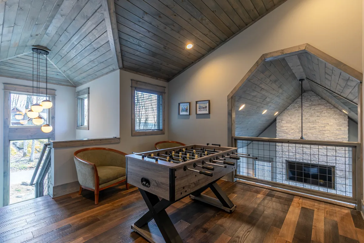 This image showcases a vibrant media lounge featuring a foosball table prominently in the center, complemented by a wooden ceiling, large windows letting in abundant natural light, and comfortable seating along the side. The inviting, playful atmosphere is ideal for family fun and friendly competition.