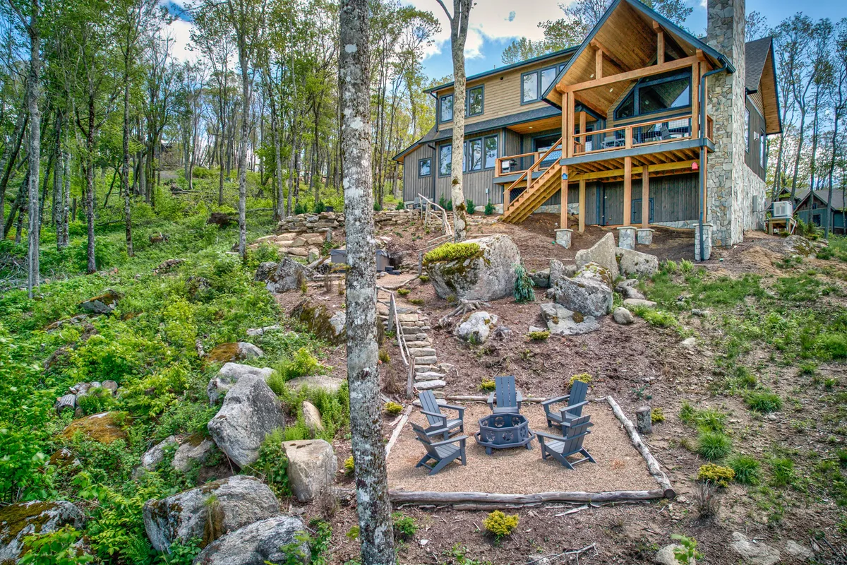 This exterior image features the house with expansive windows, a fire pit area surrounded by seating, and trees and rocks creating a natural, playful setting. The open deck is ideal for gatherings, and the fire pit evokes memories of s’mores and evening chats under the stars.