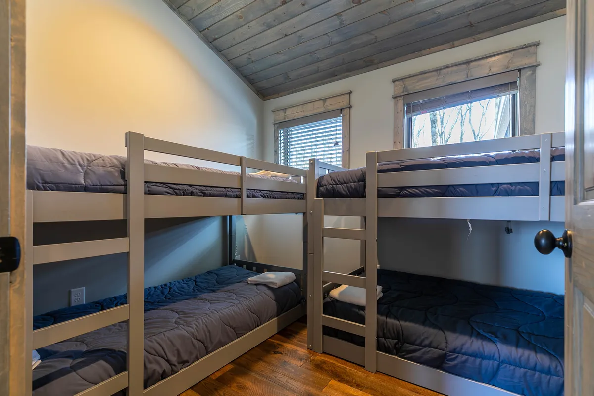 A playful yet inviting bunk room featuring two sets of sturdy bunk beds beneath a wooden ceiling. A window with blinds lets in natural light, creating a cozy space ideal for children or extra guests.