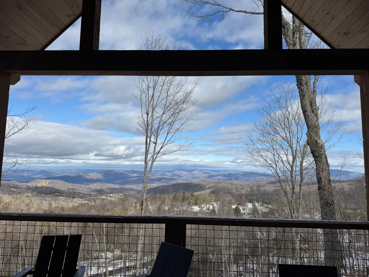 A wide view of the mountainous landscape framed by tree branches, under a blue sky. The scene captures the tranquility of the setting and hints at seasonal changes, making it ideal for activities like hiking, porch relaxation, and appreciating nature's transformation.