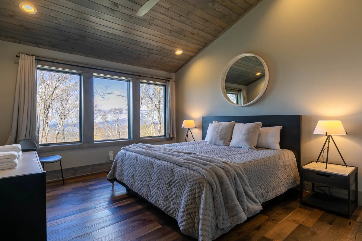 A tranquil king bedroom with large windows framing peaceful wooded or mountain views. The plush bed is topped with crisp linens and complemented by a round mirror above the headboard, inviting relaxation and a connection with the serene surroundings.