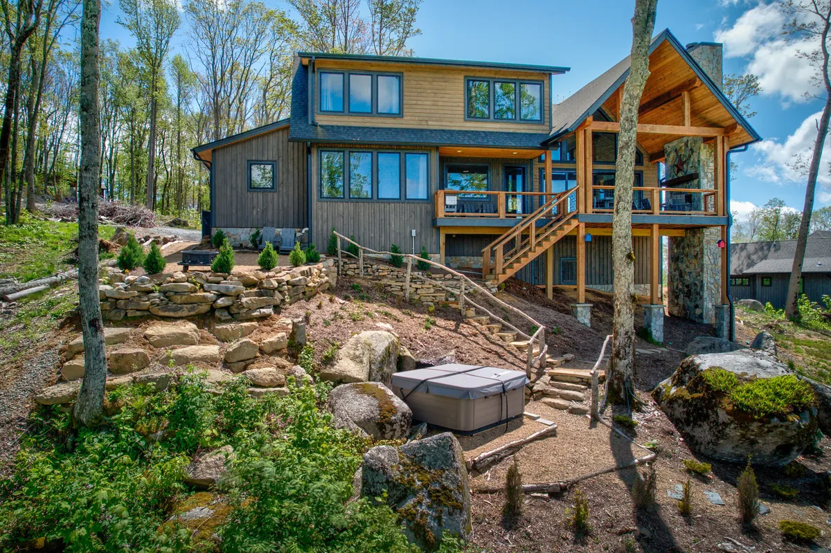 This image features a large modern house with a relaxing hot tub on a spacious deck, a stone pathway, and a beautifully landscaped garden, all surrounded by lush nature. The setting is serene, inviting, and perfect for private retreats or social gatherings.