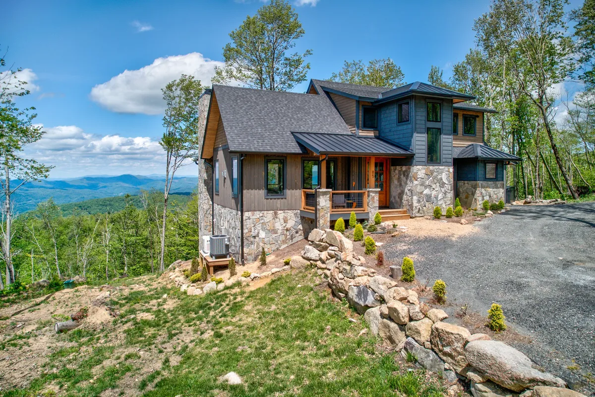 A serene exterior shot highlighting the home's modern architecture with a stone facade, lush greenery, and a breathtaking mountain backdrop. Two Adirondack chairs on the porch invite outdoor relaxation, making it easy to imagine enjoying a coffee in the morning or stargazing at night.