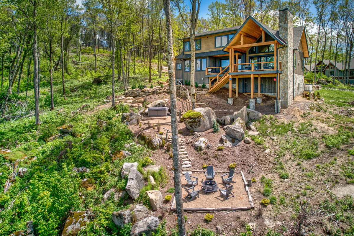 An inviting view of the house with a spacious outdoor seating area, fire pit, and surrounding trees. The scene suggests areas for gatherings, relaxation, and playful outdoor moments with pets, set against a tranquil backdrop.