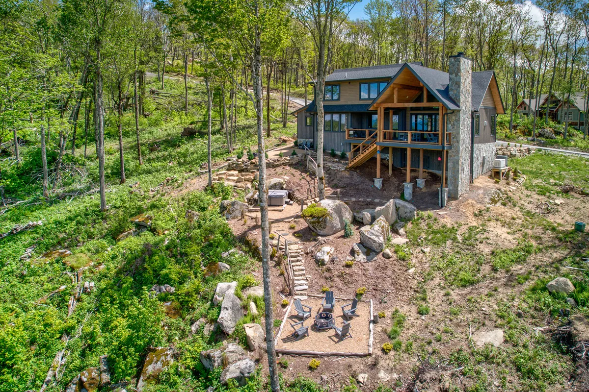 The photo showcases a modern house with a wooden deck, a dedicated fire pit area, and natural stone elements, all surrounded by lush woods. The scene hints at communal relaxation after a day of adventure, such as gathering around a fire or setting out for a hike.
