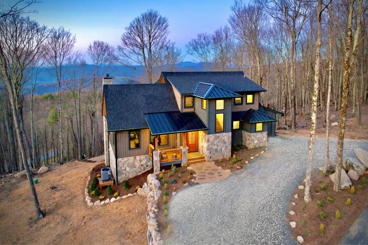 The image showcases a striking modern mountain home nestled among tall trees, with a natural stone facade, expansive windows, and an inviting driveway. Sunlight bathes the front porch, hinting at breathtaking sunset views and the tranquility of surrounding nature.