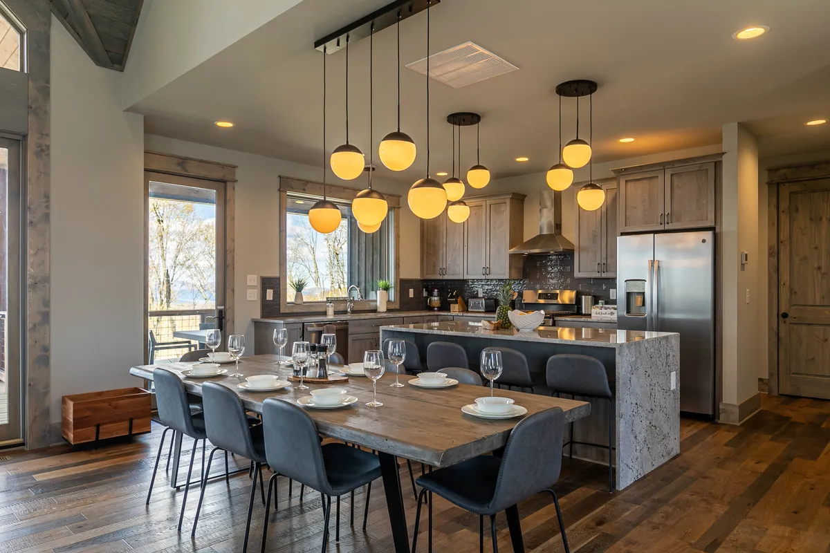 A contemporary dining area set for a meal, with unique pendant lighting and a spacious adjoining kitchen. The setup is warm, welcoming, and ready for family feasts or dinner parties, reflecting the home’s capacity for shared experiences.