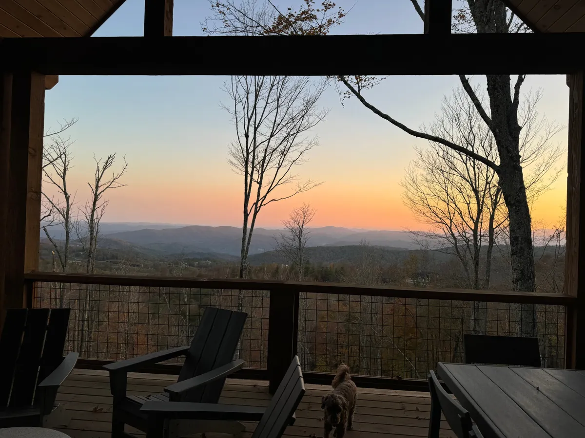 This outdoor view showcases a mountain landscape at sunset with inviting outdoor seating. The warm hues and comfortable setup suggest an ideal spot for unwinding with a glass of wine while enjoying nature and company.