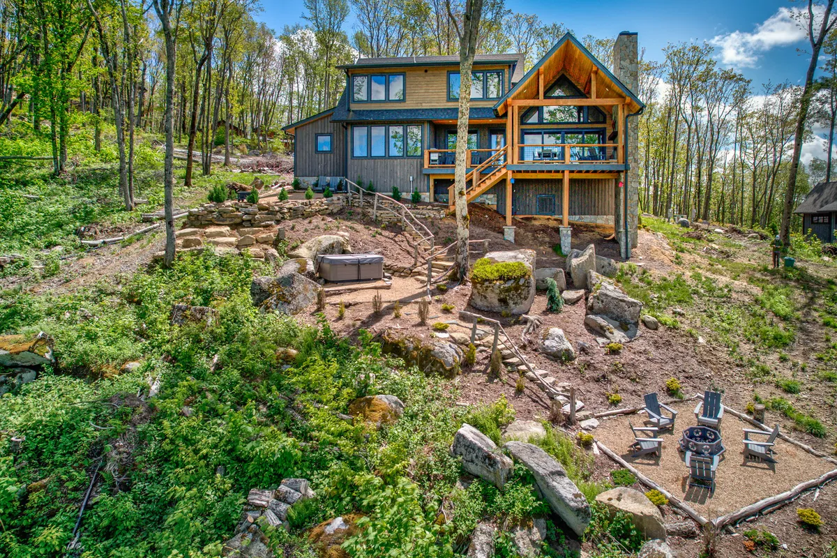 This exterior shot captures the house with large windows, an inviting deck, a dedicated fire pit area, a hot tub, and the surrounding natural mountain landscape. Both the fire pit and hot tub are clearly visible, set against a tranquil, panoramic mountain backdrop, showcasing the interconnected outdoor amenities.