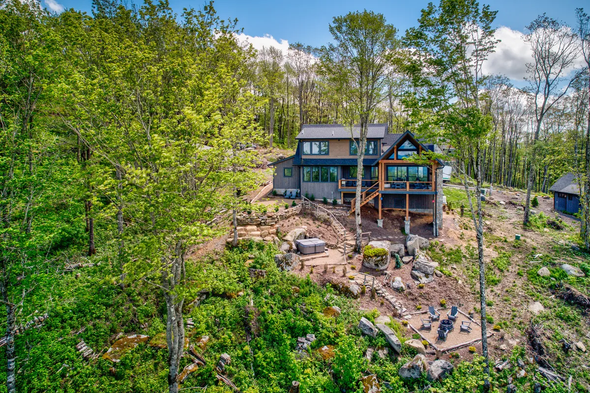 A modern home is shown with a spacious deck featuring outdoor seating, a fire pit, and views of surrounding trees and landscaped yard. The arrangement is ideal for outdoor dining, connecting with family, and soaking up the mountain atmosphere.
